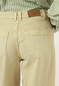 Beežid teksapüksid nahast märgisega "YAS" tagaküljel. Omadusteks on kaks tagataskut ja standardne viie taskuga disain.