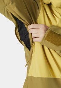 Gelbe Jacke mit einer Reißverschlusstasche und schwarzem Netzfutter. Der Stoff ist glatt mit einer matten Textur und enthält ein Klettverschluss-Patch.