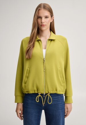 Femme aux longs cheveux raides porte une veste vert clair à fermeture éclair avec poches devant et cordons, assortie à un jean bleu, debout face à l’avant.