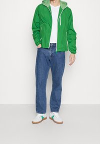 Giacca leggera verde con cappuccio verde chiaro a contrasto, abbinata a jeans in denim blu e sneakers bianche con dettagli verdi.