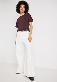 Bordo kokvilnas t-krekls apvienots ar baltām platām biksēm. Melns jostas siksna ar apļa akcentiem. Modelis nēsā tumšas saulesbrilles un melnus zābakus ar papēžiem.