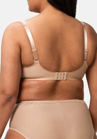 Žena oblečená v béžové podprsence a odpovídajícím vysokém spodním prádle, viděná ze zadu na jednoduchém bílém pozadí.