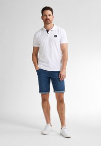 Polo bianco a maniche corte con dettagli neri al colletto, abbinato a pantaloni blu in denim. Il modello indossa sneaker bianche, posando su uno sfondo chiaro.
