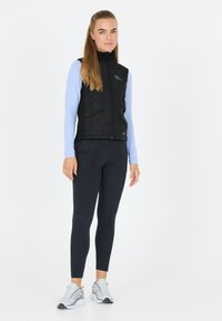 Schwarze gesteppte Weste mit hohem Kragen und Reißverschluss vorne, übereinander getragen mit einem hellblauen Langarmoberteil, kombiniert mit schwarzen Leggings und weißen Turnschuhen.