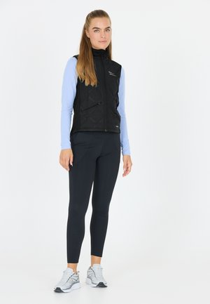 Schwarze gesteppte Weste mit hohem Kragen und Reißverschluss vorne, übereinander getragen mit einem hellblauen Langarmoberteil, kombiniert mit schwarzen Leggings und weißen Turnschuhen.