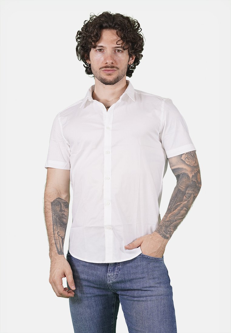 Camicia bianca a maniche corte con colletto classico, tessuto liscio e taglio dritto. Indossata con jeans blu, che mettono in mostra i tatuaggi sulle braccia.