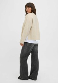 Suéter de punto acanalado beige con un corte cropped, usado sobre una camisa blanca, combinado con jeans sueltos de un gris oscuro y zapatillas sin cordones negras.