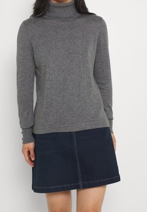 Femme portant un pull gris à col roulé à manches longues et une jupe en jean bleu foncé, debout devant un fond blanc uni.