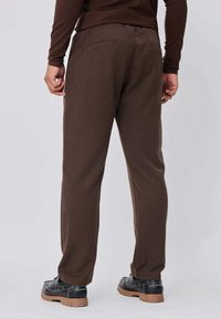 Personne portant un pantalon marron et un haut à manches longues assorti, vue de dos, debout sur un fond uni gris clair.