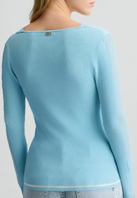 Haut bleu clair à manches longues côtelées avec un col arrière arrondi, orné d'un accent argenté à l'arrière et en tissu texturé.