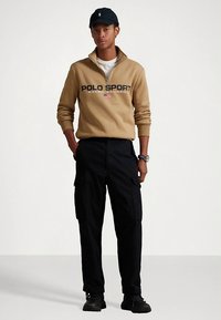 Άνδρας που φοράει ανοιχτόχρωμο μπεζ φούτερ Polo Sport με φερμουάρ μέχρι το στήθος, μαύρο cargo παντελόνι, μαύρα αθλητικά παπούτσια, μαύρο καπέλο και ρολόι, με φόντο απλό και φωτεινό.