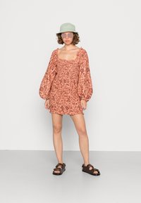 Vestido mini com padrão floral em pêssego, com mangas bufantes e corpete franzido. Usado com um chapéu de balde verde claro e sandálias escuras.