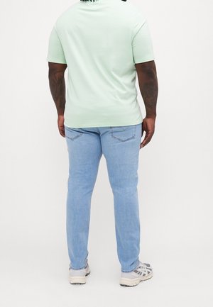 Mann trägt hellgrünes Kurzarmhemd, hellblaue Jeans und graue Sneakers, steht mit dem Rücken zu einem schlichten weißen Hintergrund.