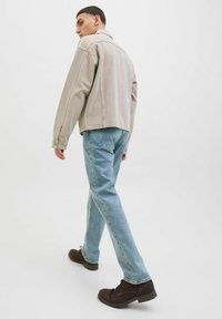 Jachetă din denim bej cu umeri lăsați, blugi drepți de culoare bleu deschis și pantofi maronii, văzută din spate pe un fundal alb.