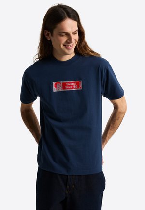 Joven con cabello largo sonriendo, vistiendo una camiseta azul marino con un parche del logo de Vans en rojo y blanco que dice "Sickest Since '66."