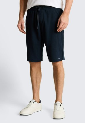 Mann mit schwarzen Kordelzug-Shorts und weißen Sneakers steht vor einem schlichten hellen Hintergrund.