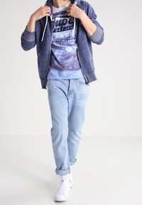 Blauer Kapuzenpullover mit Reißverschluss, hellblaues grafisches T-Shirt, Jeans in heller Waschung, weiße Sneaker. Lässiges Outfit mit entspannter Passform und aufgekrempelten Hosenbeinen.