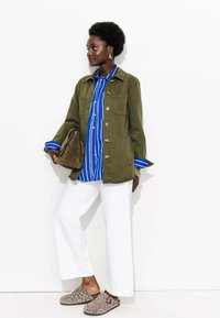 Femme portant une veste verte, une chemise à rayures bleues, un pantalon large blanc, des chaussures à enfiler à imprimé léopard et tenant un sac à main en daim marron.