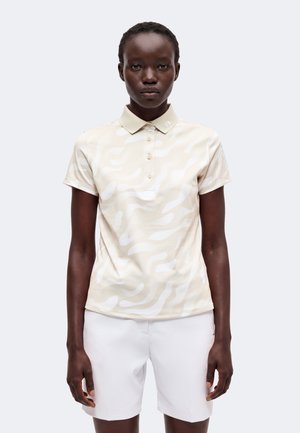 Junge schwarze Frau trägt ein beige gemustertes kurzärmeliges Polohemd und weiße Shorts, steht vor einem einfarbigen hellen Hintergrund.