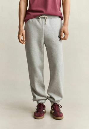 Man draagt lichtgrijze joggingbroek met trekkoord, een rood shirt en bordeauxrode en beige sneakers, staand op een effen vloer.