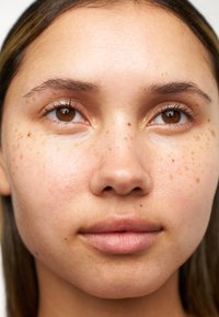 Close-up van een gezicht met een gladde, lichtbruine huid, opvallende sproeten, bruine ogen en zachte, natuurlijke lippen. Het haar is steil en lichtgekleurd.