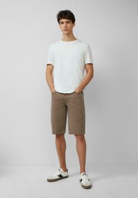 Camiseta blanca de algodón, pantalones cortos marrones hasta la rodilla y zapatillas con detalles en negro. El modelo está de pie con las manos en los bolsillos frente a un fondo liso.