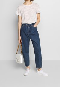 Biała bluzka z krótkim rękawem z drobnym kwiatowym wzorem, zestawiona z niebieskimi spodniami dżinsowymi o wysokim stanie i białymi trampkami, trzymająca białą torbę.