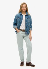 Chaqueta de mezclilla en azul, camiseta blanca, pantalones verde claros y zapatos marrones con cordones. La chaqueta tiene bolsillos y mangas remangadas.