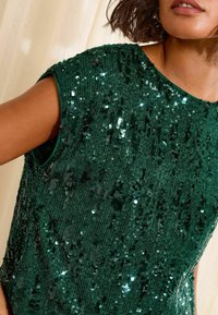 Top verde ricoperto di paillettes, caratterizzato da un tessuto testurizzato con scollatura rotonda e maniche corte. Le paillettes sono disposte densamente, aggiungendo lucentezza.
