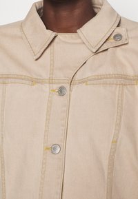 Veste en denim beige avec un col, des détails en métal argenté, un design structuré et des accents de couture jaune le long des coutures.