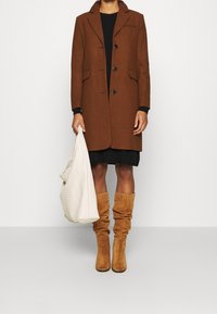 Brun uldfrakke med tre knapper, spidse krave; parret med en sort kjole, en beige tekstureret taske og brune knæhøje slouch støvler.