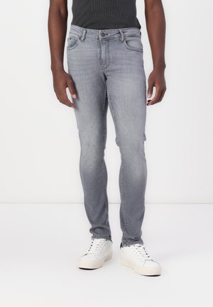 Homme portant un jean slim gris délavé, des baskets blanches et un haut noir sans manches, debout devant un fond blanc uni.