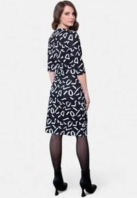 Robe noire avec des formes abstraites blanches, manches trois-quarts, taille cintrée et longueur au genou. Portée avec des bottes à talons hauts noires.