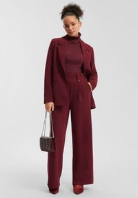 Completo bordeaux con un blazer su misura e pantaloni a gamba larga, abbinato a un dolcevita coordinato e a una piccola borsa decorata.