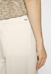 Harmaat beige-housut, jotka on valmistettu sileästä kankaasta. Niissä on takatasku, jossa on hienovarainen logo-detaili ja siistit ompeleet saumoissa.