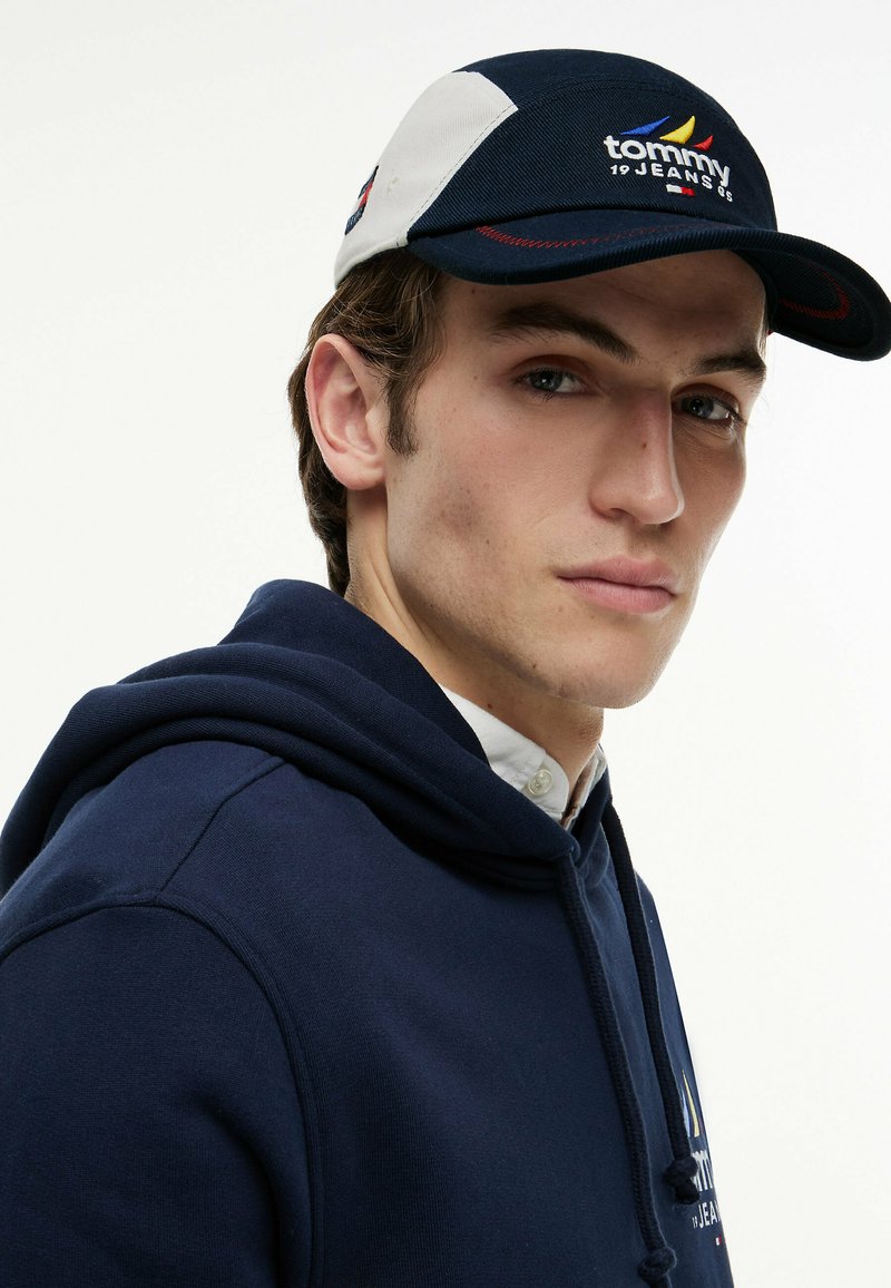 Young man wearing a navy blue and white Tommy Jeans cap and navy blue hoodie, facing partially sideways against a white background.