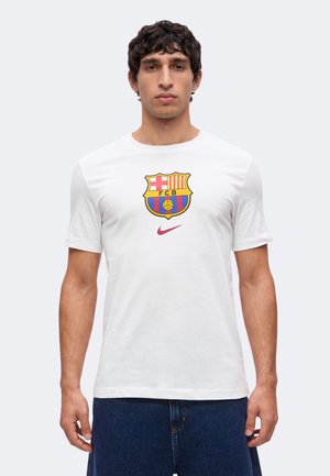 Homme portant un t-shirt blanc avec le logo du FC Barcelone et le swoosh rouge Nike, associé à un jean bleu foncé, debout devant un fond clair uni.