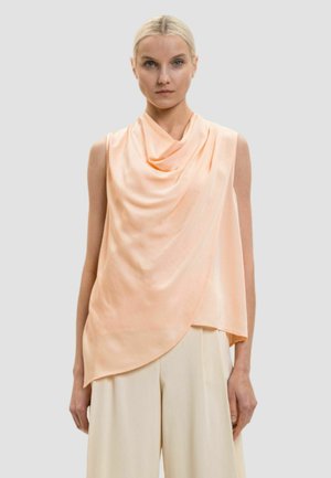 Donna che indossa una blusa drappeggiata color pesca senza maniche abbinata a pantaloni crema ampi, in piedi davanti a uno sfondo grigio semplice.