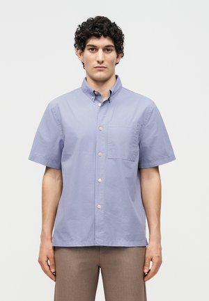 Άνδρας με σγουρά σκούρα μαλλιά, φοράει ένα ανοιχτό μπλε πουκάμισο με κοντά μανίκια και καφέ παντελόνια, στέκεται μπροστά σε ένα απλό φόντο.