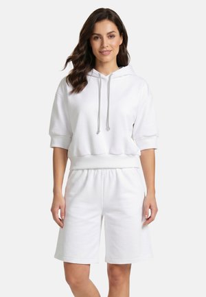 Vrouw met los golvend haar, gekleed in een witte korte mouw hoodie en bijpassende losse witte korte broek tegen een witte achtergrond.