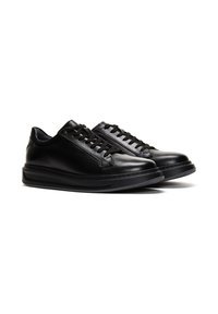 Para czarnych skórzanych sneakersów typu low-top z czarnymi sznurowadłami i grubymi czarnymi gumowymi podeszwami na białym tle.