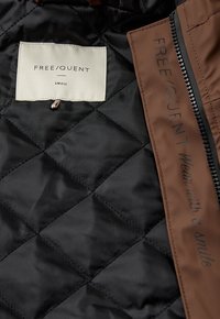 Zwarte gewatteerde binnenkant van een jack met een crème label waarop staat "FREE/QUENT SMALL" en een bruine contrasterende rits met bedrukte tekst.