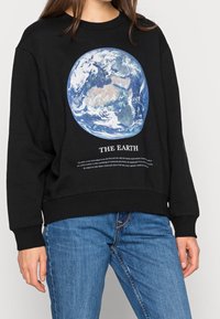 Personne portant un sweat-shirt noir avec un graphisme de la Terre et le texte « THE EARTH », associée à un jean bleu, debout devant un fond blanc.