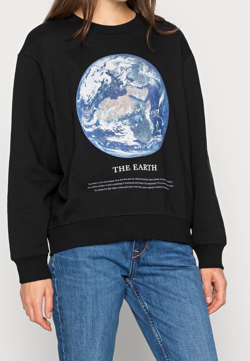 Personne portant un sweat-shirt noir avec un graphisme de la Terre et le texte « THE EARTH », associée à un jean bleu, debout devant un fond blanc.