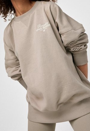 Sweatshirt beige en tissu doux, doté de manches tombantes, de poignets côtelés et d'une broderie "Saison de la cannelle" à l'avant.