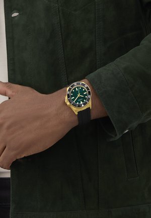 Orologio dorato con quadrante verde, cinturino in silicone nero e lunetta girevole. Il display presenta indici delle ore e tre lancette.