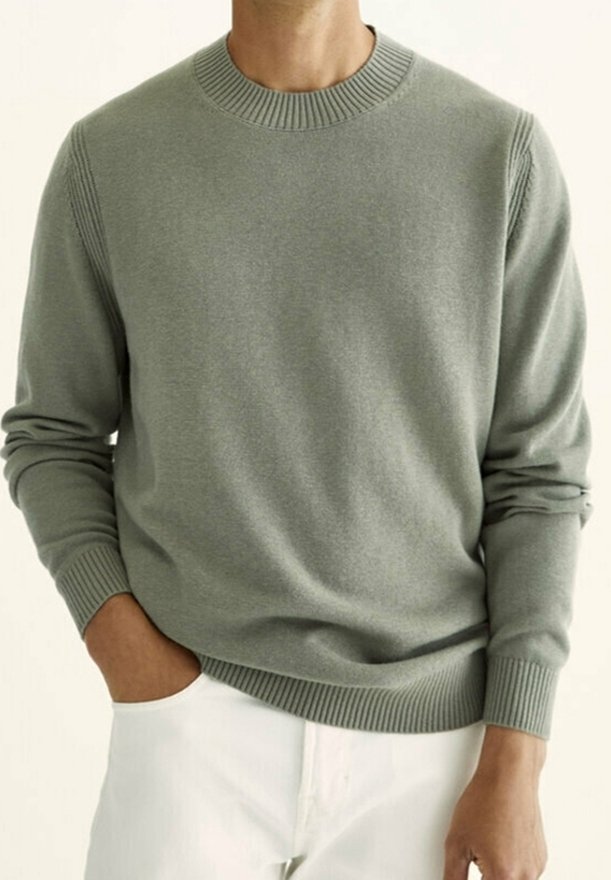 Homme portant un pull gris clair à col rond côtelé à manches longues et un pantalon blanc, une main dans la poche et l'autre détendue le long du corps.