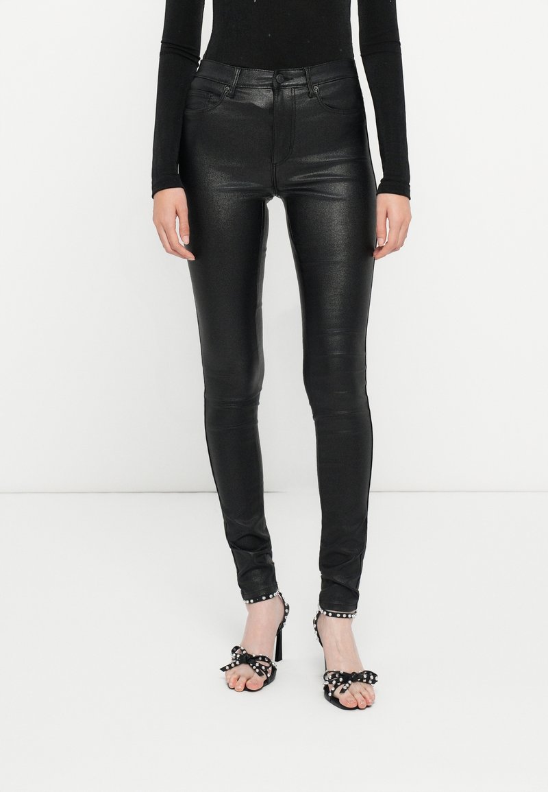 Personne portant un pantalon en cuir noir brillant taille haute et des sandales à talons hauts noires ornées de nœuds cloutés de perles sur un fond blanc.
