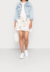 Veste en jean sur un t-shirt clair, associée à une jupe blanche à motif floral et des baskets blanches. Accessoires comprenant un sac blanc texturé.