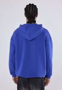 Person mit geflochtenem Haar, der einen königsblauen Kapuzenpullover und ausgewaschene schwarze Jeans trägt, steht vor einem schlichten hellen Hintergrund mit dem Rücken zugewandt.